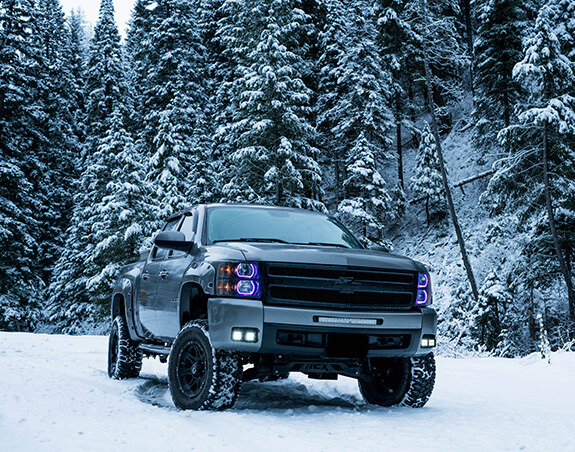 Old Chevy Silverado driving through the snow with ever green tress in the background